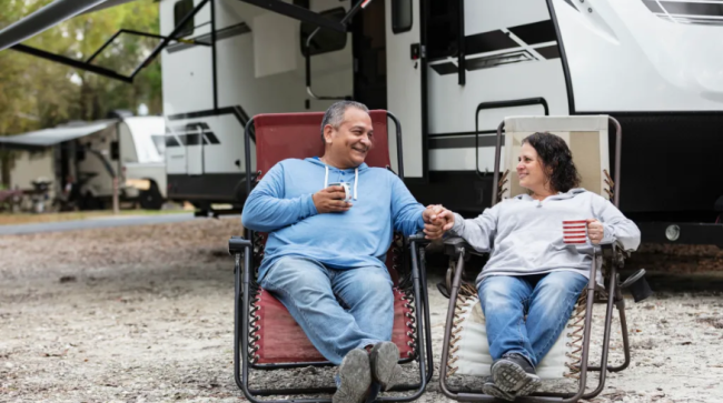 A couple sitting together in chairs by a camper trailer in an RV park, drinking coffee.