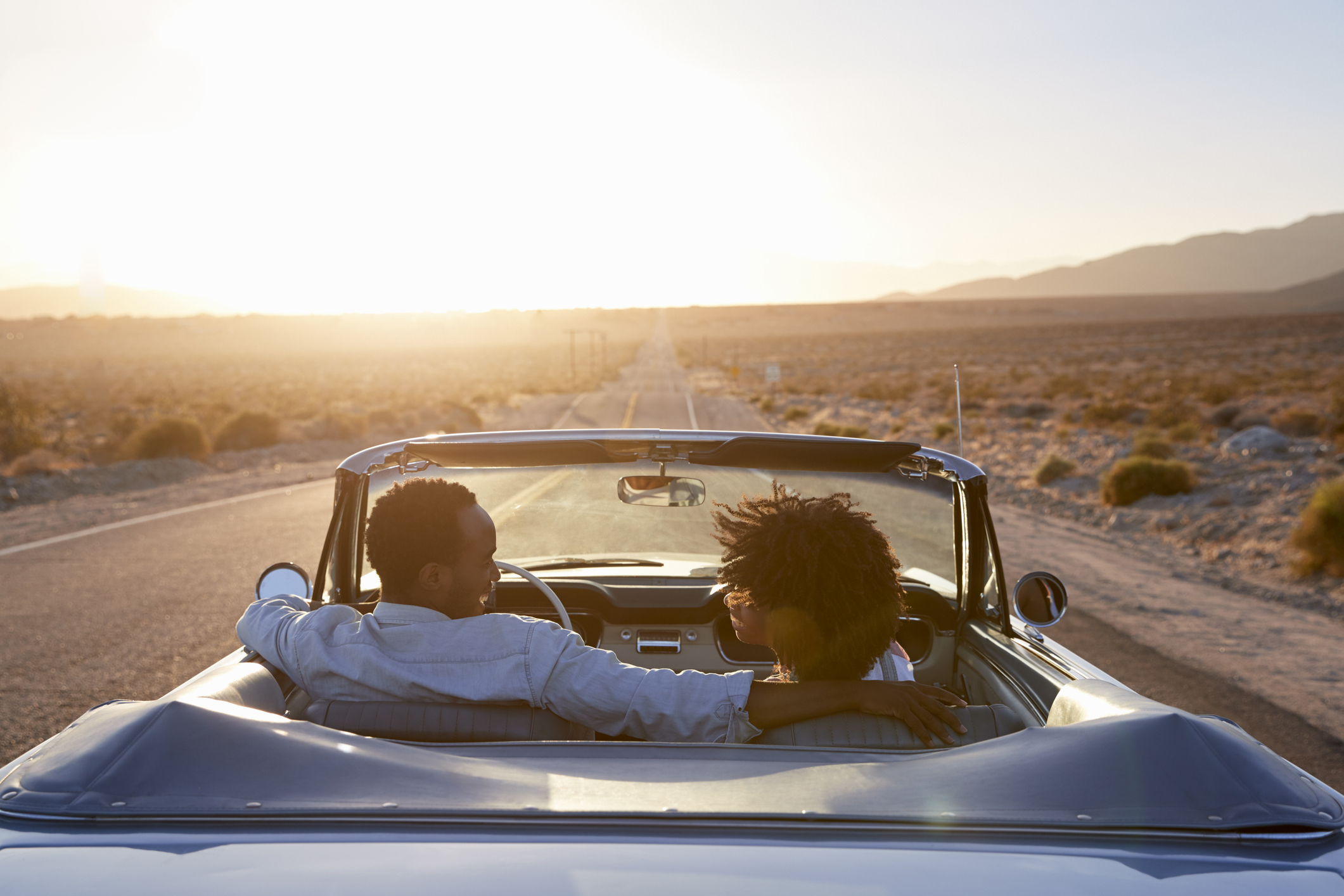 Rear view of couple on road trip driving classic convertible car towards sunset.