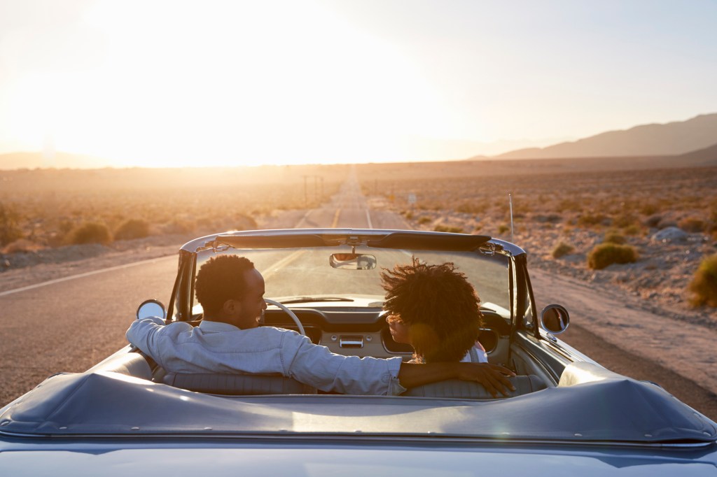 Rear view of couple on road trip driving classic convertible car towards sunset.