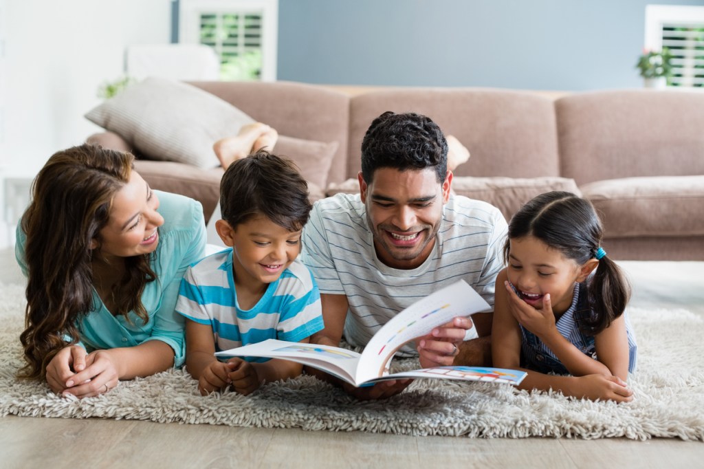 Parents and children lying on rug and reading book in living room at home.