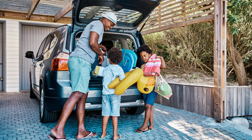 Dad and two kids loading a teal SUV with beach gear from a driveway.